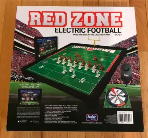 An electric football board game titled "RED ZONE ELECTRIC FOOTBALL" with miniature players set up on a miniature field, a handheld remote control, and packaging that lists game features and contents.