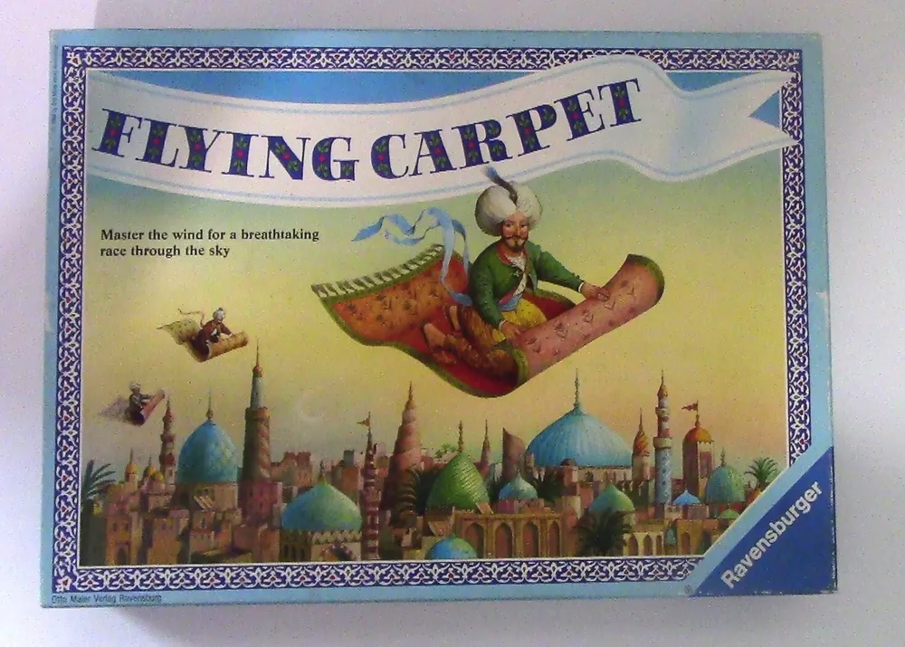The image is of a board game box titled "FLYING CARPET" by Ravensburger, featuring artwork of characters riding flying carpets above a city with blue and green domed buildings.