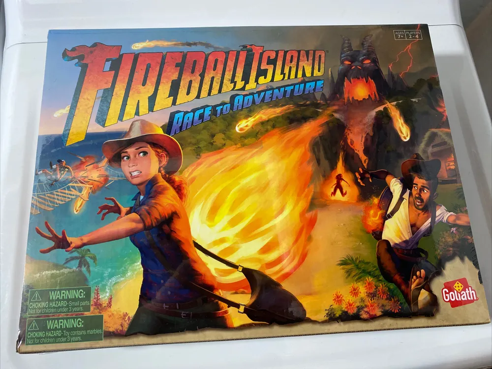 The image shows a board game cover titled "FIREBALL ISLAND: RACE TO ADVENTURE". Illustrated on the cover is a scene with two characters, a man and a woman, both wearing hats and looking panicked as they flee from a fireball. In the background, there is a large, fiery Tiki statue and a bridge with people running across it. The box includes warning labels about choking hazards and the Goliath company logo.