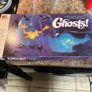 A worn board game box labeled "Ghosts!" with a tagline "A VERY CREEPY, SNEAKY Guess-Who Game" is displayed on a kitchen counter. The box art features cartoonish ghosts and spiders against a purple backdrop, indicating the spooky theme of the game. The upper left corner shows the MB (Milton Bradley) logo and text stating it's for ages 6-14 and 2 players.