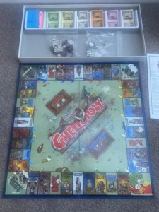 A board game called "Ghettopoly" laid out on the floor with cartoonish street-themed artwork, various card decks, and money arranged in a box insert, alongside houses, dice, and game instructions.