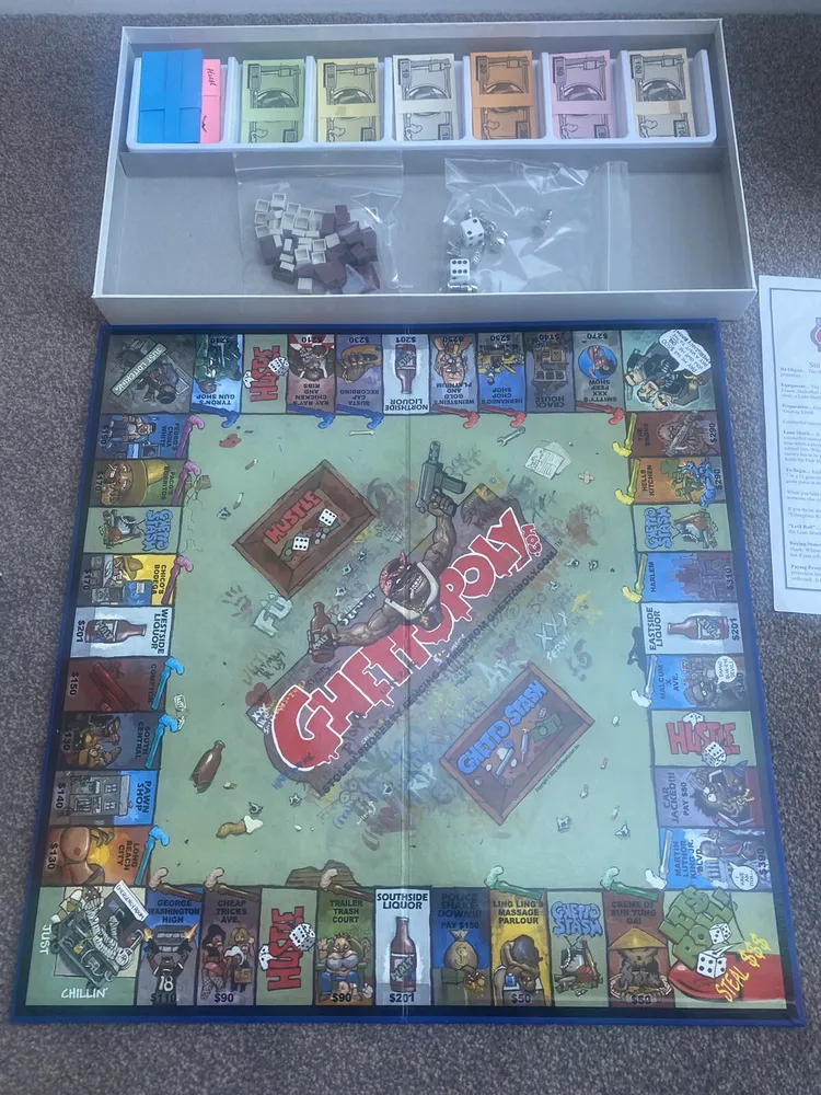 A board game called "Ghettopoly" laid out on the floor with cartoonish street-themed artwork, various card decks, and money arranged in a box insert, alongside houses, dice, and game instructions.