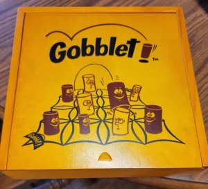 A yellow box with the text "Gobblet" and cartoon illustrations of smiling cylindrical characters, resembling game pieces, on a spider web pattern.