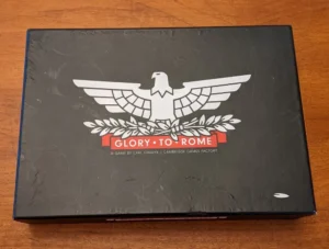 Black board game box with the title "Glory to Rome", featuring a white eagle above laurel branches, with the text "A GAME BY CARL CHUDYK / CAMBRIDGE GAMES FACTORY" underneath. The box is lying on a wooden surface.