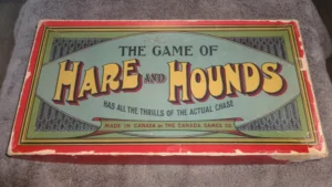 Vintage board game box titled "The Game of HARE AND HOUNDS" with a tagline "HAS ALL THE THRILLS OF THE ACTUAL CHASE," made in Canada by the Canada Games Co., laid out on a grey fabric surface.