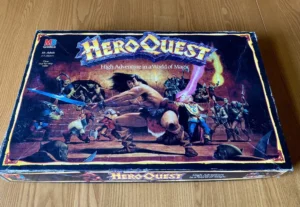 The image shows a "HeroQuest" board game box with vibrant fantasy artwork depicting various heroes and creatures engaged in battle, with the game's subtitle "High Adventure in a World of Magic" visible.