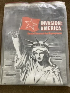 A plastic-wrapped board game box titled "Invasion: America" with the subtitle "Death Throes of the Superpower" featuring an illustration of the Statue of Liberty and a raised red flag with a star.