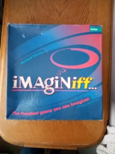 A board game named "Imaginiff..." with the tagline "The funniest game you can imagine!" for ages 10 & up, 3-8 players. The box is a revised edition by Buffalo Games with a blue and pink color scheme. The game is on a wood surface with partial view of a white object in the bottom right corner.