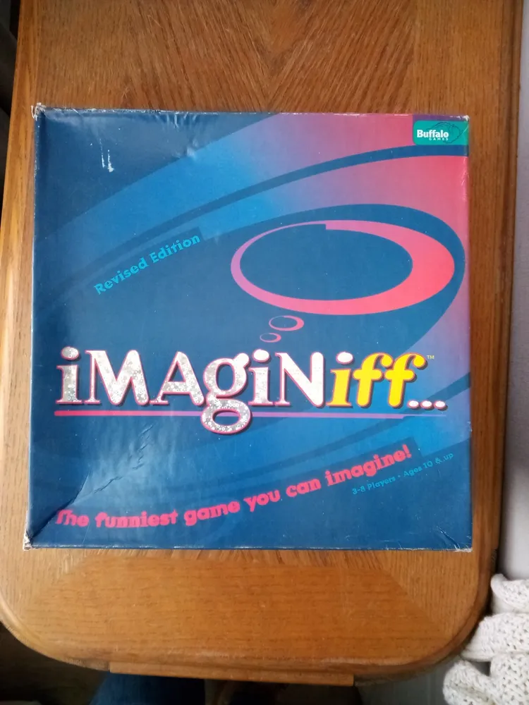 A board game named "Imaginiff..." with the tagline "The funniest game you can imagine!" for ages 10 & up, 3-8 players. The box is a revised edition by Buffalo Games with a blue and pink color scheme. The game is on a wood surface with partial view of a white object in the bottom right corner.