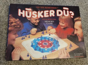 A family playing the "Husker Du?" memory game, with a mother, father, son, and daughter smiling and engaged around the game board. The box reads "Do You Remember? Husker Du? Match-the-Pictures Memory Game" for 2 or more players aged 5 and up, with instructions in Spanish included.