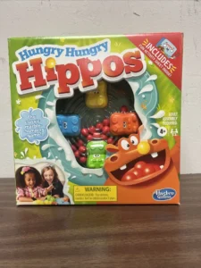 A boxed Hungry Hungry Hippos game by Hasbro Gaming, featuring colorful graphics of hippos and marbles, with an age recommendation of 4+ and a choking hazard warning. Two children are depicted playing the game on the bottom left corner.