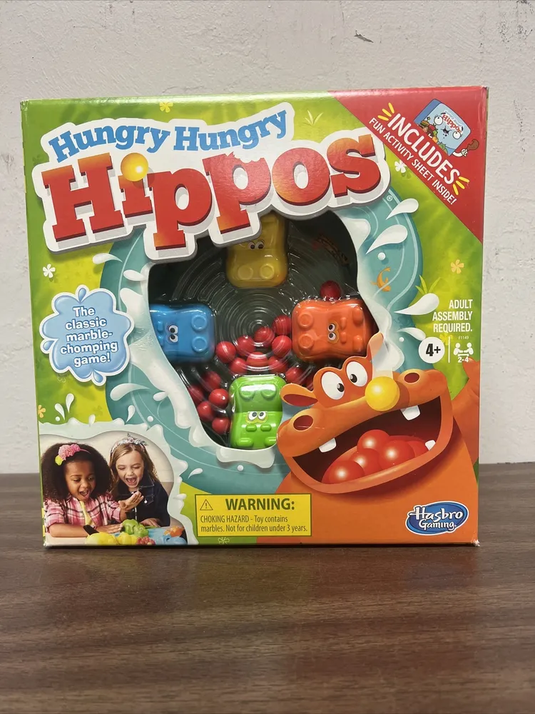 A boxed Hungry Hungry Hippos game by Hasbro Gaming, featuring colorful graphics of hippos and marbles, with an age recommendation of 4+ and a choking hazard warning. Two children are depicted playing the game on the bottom left corner.