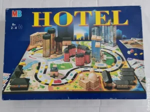 Board game "HOTEL" by MB with a recommended age of 8+ for 2-4 players, displaying the game's cover with colorful graphics of buildings, a winding road, a game plan, and game components.