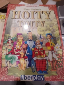 Box cover of the board game "Hoity Toity" showing illustrated characters, including a woman with a green dress and hat, a man with a monocle and teddy bear, a man in a wheelchair with medals, and a man with a polo mallet, all depicted in a playful, aristocratic style against the backdrop of a mansion.