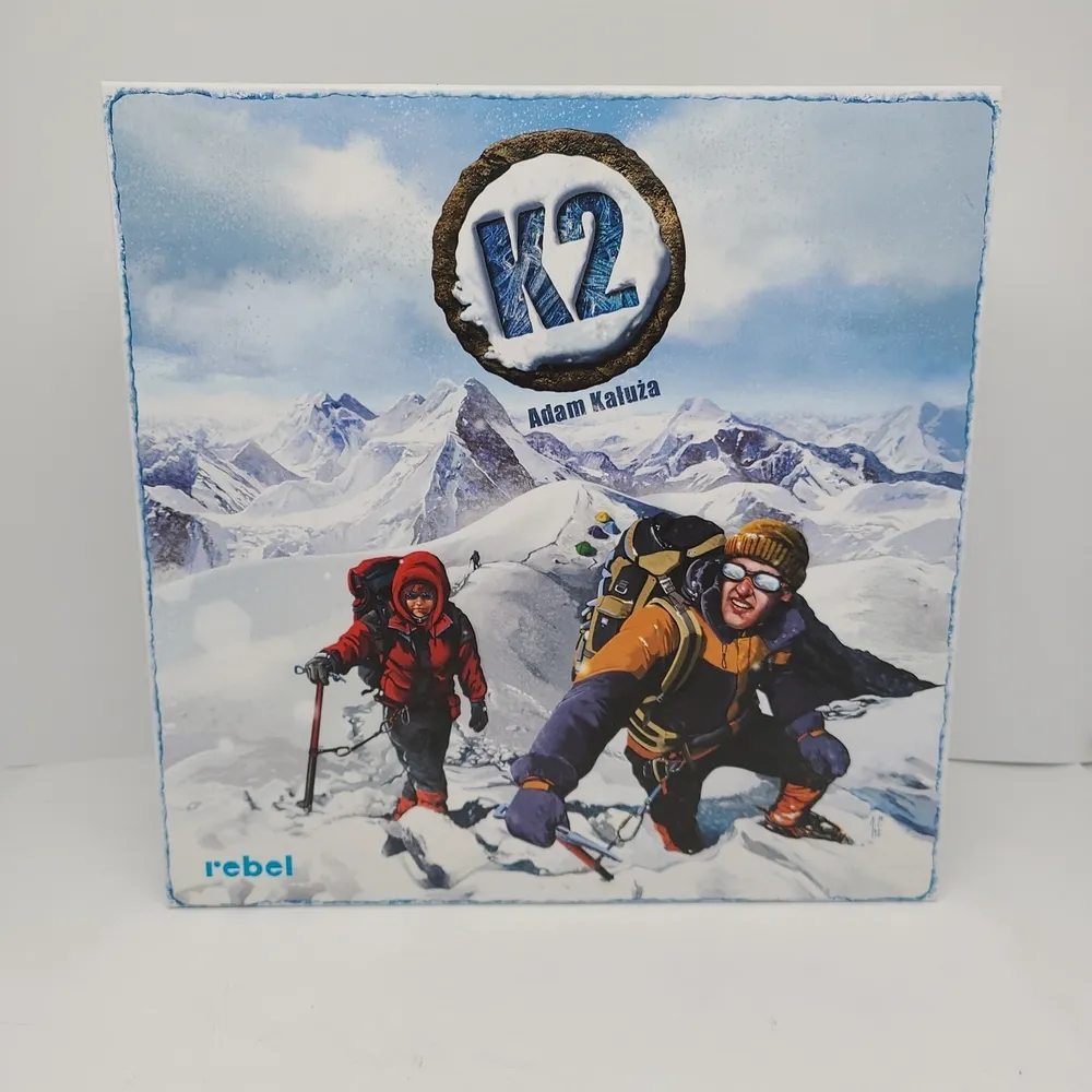 Board game cover featuring two climbers in heavy gear on a snowy mountain slope with the game title "K2" and the name "Adam Kałuża" above, and the logo of "rebel" at the bottom.