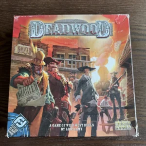 A board game named "Deadwood" on a wooden table, featuring an illustration of Wild West characters engaged in a gunfight, with the subtitle "A Game of Wild West Duels" by Loïc Lamy and logos for "Pretzel Games" and "Dust Games" on the box.