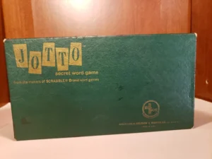 A vintage "JOTTO secret word game" box with worn green cover, gold lettering, and the SCRABBLE brand logo, manufactured by Selchow & Righter Co., displayed against a wooden backdrop.
