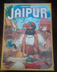 A board game named "JAIPUR" with an illustration of a bearded man in traditional Indian attire holding a blue pot, sitting beside trade goods and a camel, with Indian architecture in the background.