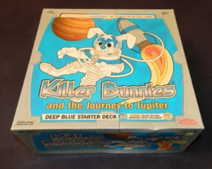 A boxed board game titled "Killer Bunnies and the Journey to Jupiter" featuring a cartoon bunny astronaut, with text indicating it's the "Deep Blue Starter Deck" and includes a "Solar Yellow Booster Deck." The box lies on a dark surface.