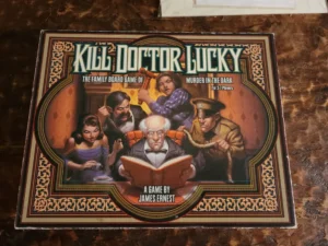 A "Kill Doctor Lucky" board game box depicting cartoon-style characters -- a young woman, two men, an elderly man, and a person in the shadows -- seated around a room, with the elderly man reading a book, oblivious to the others' malicious intentions. The cover also includes the subtitle "Murder in the Dark" and text indicating it is for 3-7 players and designed by James Ernest.