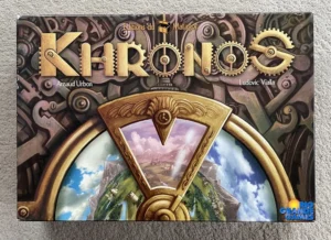 A board game cover for "KAIROs" with a steampunk-themed background featuring gears and cogs, displayed on a textured surface. The title is spread across the top with the names Arnaud Urbon and Ludovic Vialla below it, and the logos for Editions du Matagot and Rio Grande Games are visible on the sides. The central illustration shows a golden-rimmed hourglass with a picturesque landscape view through its upper half.