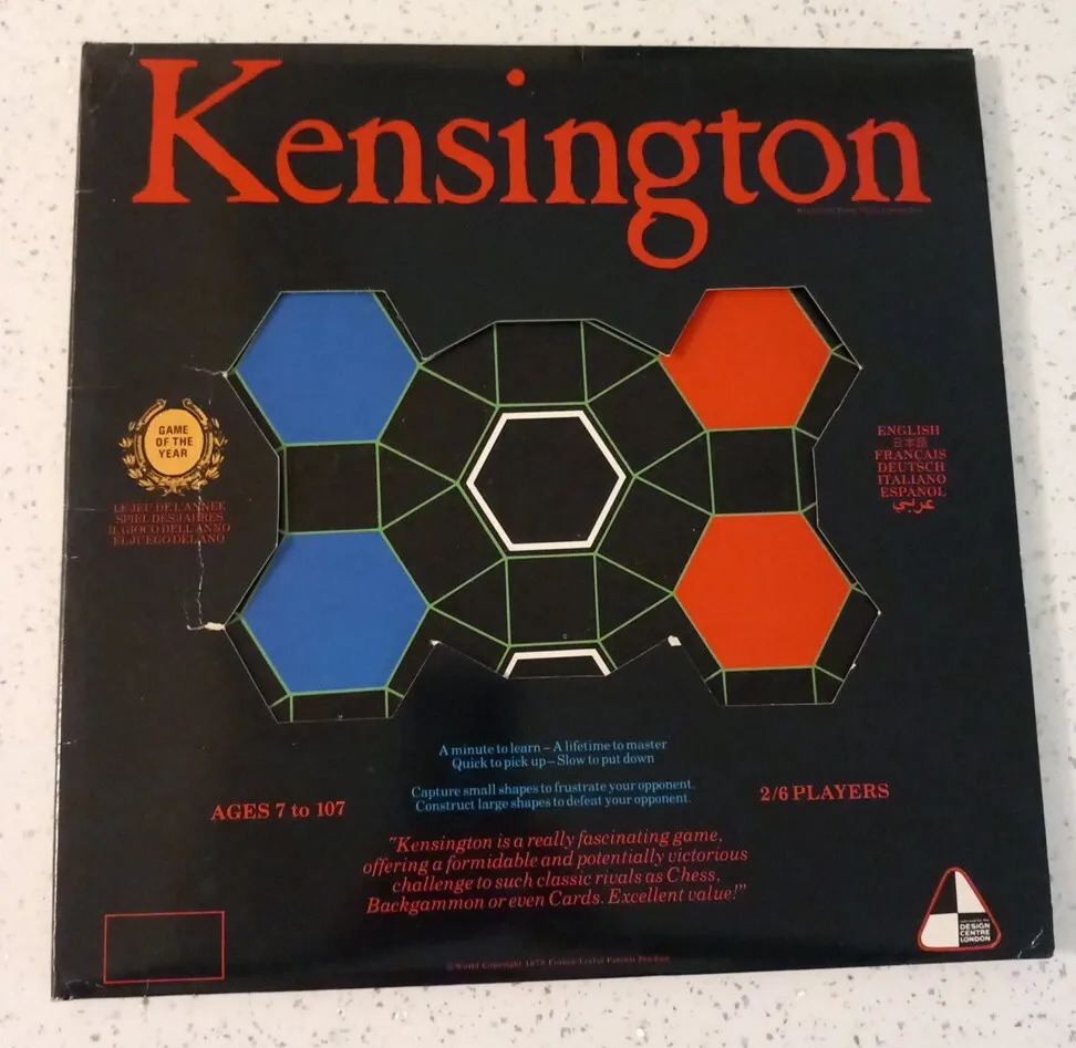 Box cover for "Kensington" board game, featuring hexagonal patterns with game title in large red letters, subtitle text, age range (7 to 107), player number (2/6 players), and multilingual instructions. A sticker on the corner indicates it was the "Game of the Year."