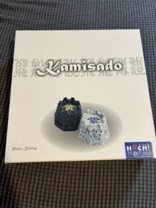 A board game box for "Kamisado" by Peter Burley with two game pieces on top, one black and one white, both featuring Asian characters, and a "HUCH & friends" logo on the bottom right corner, all placed on a grey woven surface.