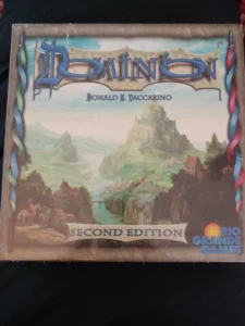 Box cover of the "Dominion" Second Edition board game by Donald X. Vaccarino, published by Rio Grande Games, featuring a medieval landscape with a castle on a hill.