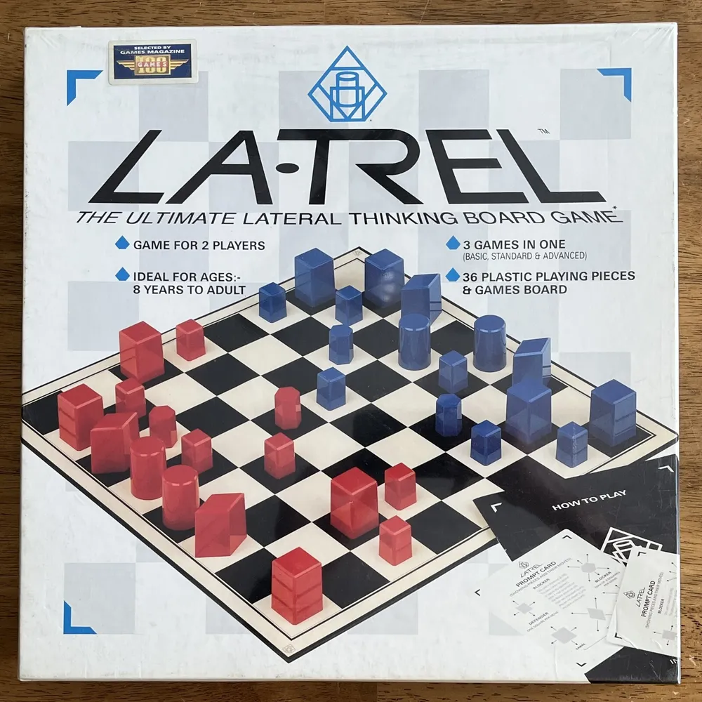 A board game called 'LA-TREL The Ultimate Lateral Thinking Board Game' is displayed on a table, with a chessboard-like pattern containing red and blue plastic pieces, ideal for 2 players and designed for ages 8 to adult, including a rule sheet with instructions on how to play.