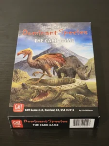 A photo of the board game "Dominant Species: The Card Game" by Chad Jensen, with illustrated dinosaurs and ancient animals on the cover. The game is published by GMT Games and displays their logo along with a spine label, barcode, and the year 2012 on the black table surface.