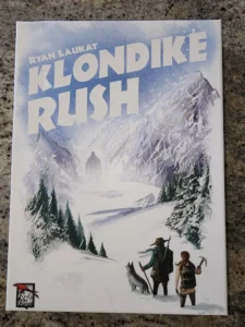 "Cover of 'Klondike Rush' board game by Ryan Laukat, featuring an illustrated snowy mountainous landscape with two adventurers and a wolf, under a title banner with the game's name, and the Red Raven Games logo in the bottom left corner."
