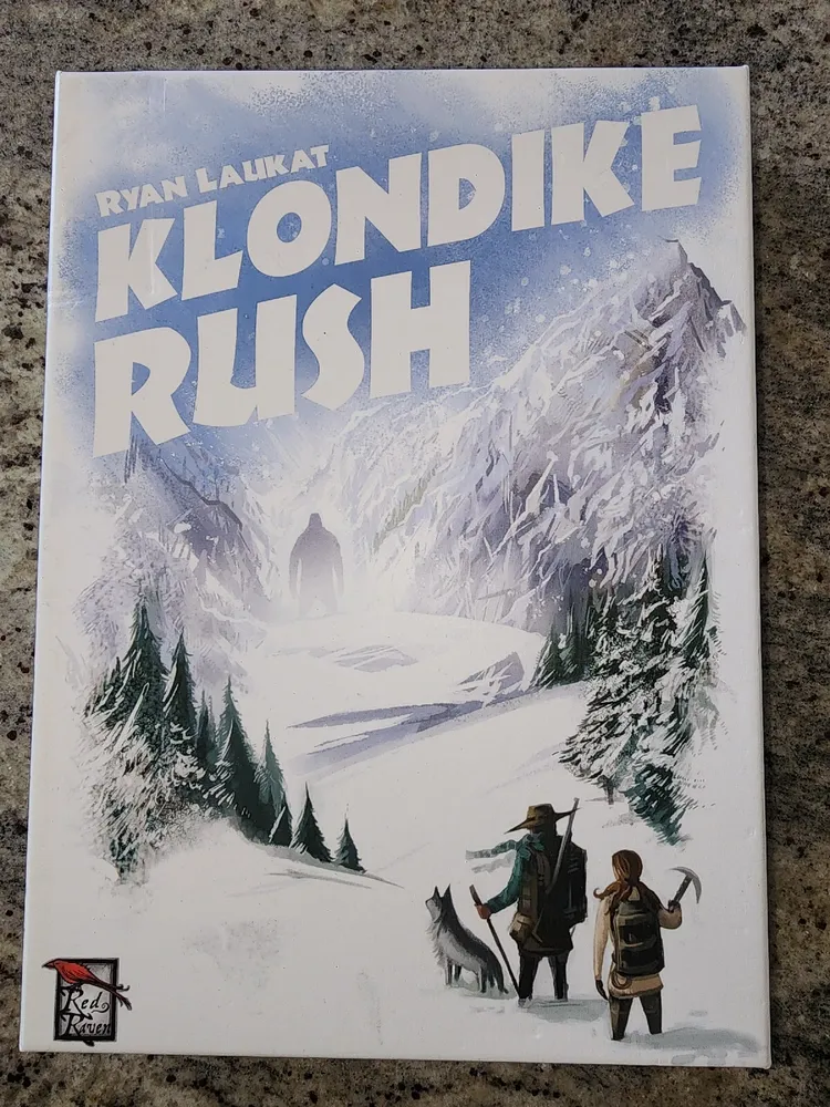 "Cover of 'Klondike Rush' board game by Ryan Laukat, featuring an illustrated snowy mountainous landscape with two adventurers and a wolf, under a title banner with the game's name, and the Red Raven Games logo in the bottom left corner."