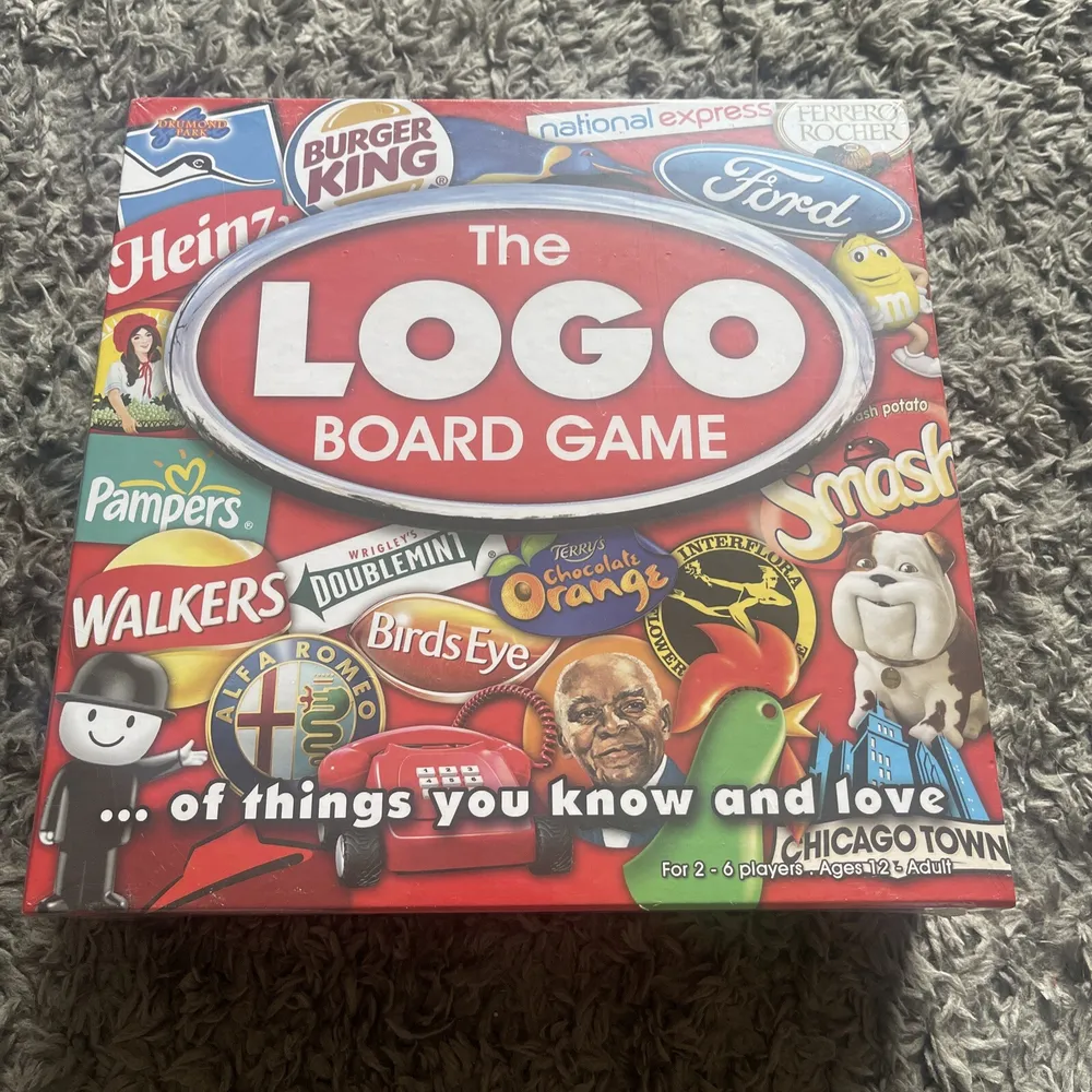 A board game box labeled "The LOGO BOARD GAME" featuring a collage of various brand logos and mascots, with the subtitle "...of things you know and love" for 2-6 players, ages 12 to adult, placed on a textured gray surface.