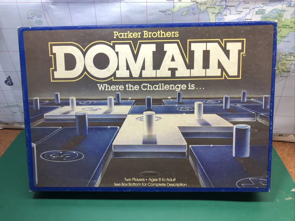 A worn boxed board game "DOMAIN" by Parker Brothers on a table with a partly visible world map in the background. The box shows a 3D illustration of abstract blue and white game components and the text "Where the Challenge is..." along with the note "Two Players - Ages 8 to Adult. See Box Bottom for Complete Description."