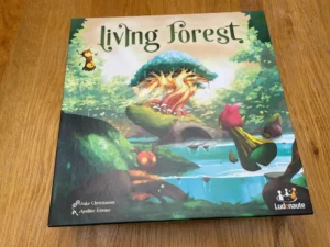 A board game titled "Living Forest" is lying on a wooden surface. The box cover features a vibrant illustration of a mystical forest with an anthropomorphic tree in the center, fire inside it, and whimsical creatures around, including a bird and small insects. The names Aske Christiansen and Apolline Étienne are credited on the box, along with the logo of Ludonaute, the publisher.