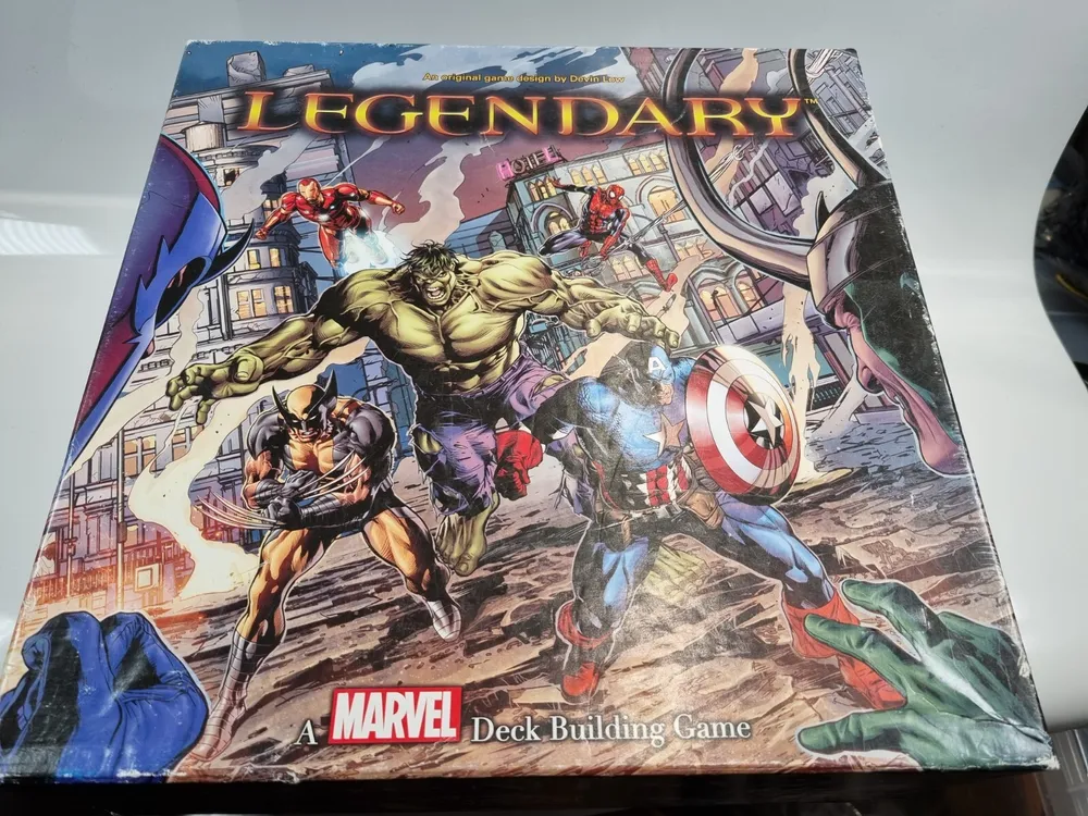 A "Legendary: A Marvel Deck Building Game" board game box featuring illustrated characters, including Wolverine, Hulk, and Captain America, among others, against an urban backdrop.