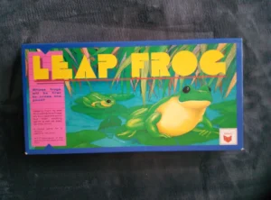 A "Leap Frog" board game box with colorful graphics of frogs on lily pads and reeds in the background. Text on the box asks, "Whose frogs will be first to cross the pond?" and notes the game is for ages 6 to 10 years. The game's manufacturer logo is visible on the lower right.