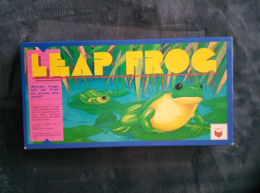 A "Leap Frog" board game box with colorful graphics of frogs on lily pads and reeds in the background. Text on the box asks, "Whose frogs will be first to cross the pond?" and notes the game is for ages 6 to 10 years. The game's manufacturer logo is visible on the lower right.