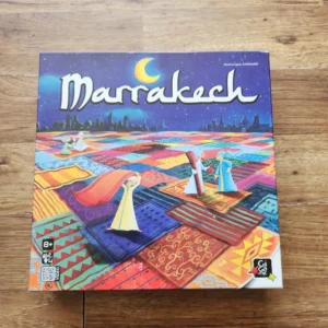 A board game named "Marrakech" with colorful, patterned artwork depicting a vibrant market scene with merchants and fabric-covered stalls, displayed on a wooden surface. The game is for ages 8 and up, created by Dominique Ehrhard, with various logos indicating publishers or certifications on the bottom left corner.