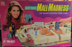 Box cover of the "ELECTRONIC MALL MADNESS" board game featuring an illustrated shopping mall with players and a large headshot of a smiling girl, with text highlighting that the game talks and is for ages 9 and up, for 2 to 4 players.