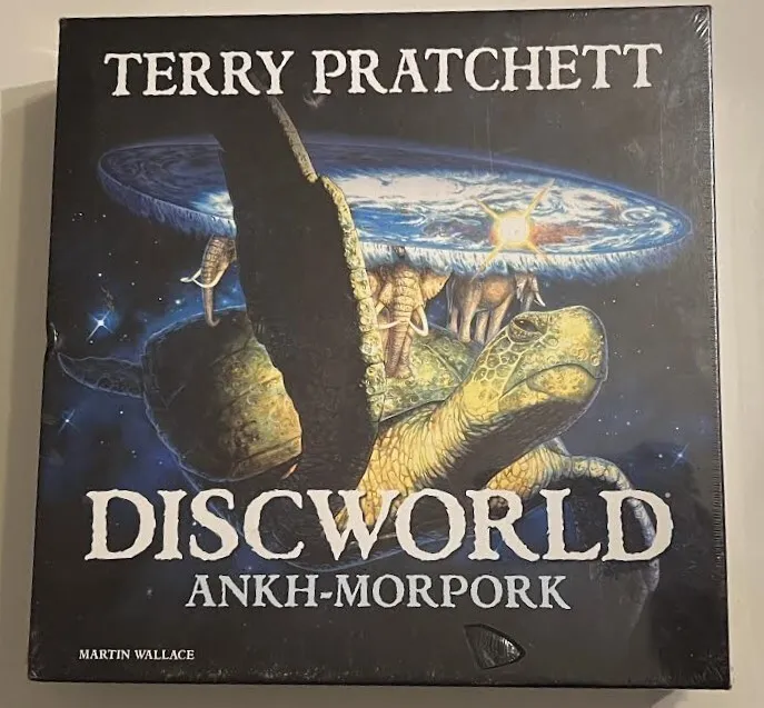 A board game cover titled "Terry Pratchett's Discworld Ankh-Morpork" designed by Martin Wallace, featuring a large turtle in space supporting a disc-shaped world with a cityscape, all against a starry background.