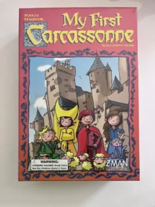 A board game box titled "My First Carcassonne" by authors Marco Teubner and Klaus-Jürgen Wrede, featuring cartoon illustrations of children in medieval costumes with a background image of castle towers and sheep, with a warning label about choking hazard for children under 3 years old.