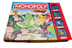 A box of "Monopoly Junior" board game by Hasbro Gaming, intended for ages 5 and up with a warning for a choking hazard. The cover features colorful cartoon illustrations of a dinosaur, a puffin, a dog, and other game pieces, alongside small windows displaying the actual game tokens: Little Scottie, Little T-Rex, Little Penguin, and Toy Ducky.