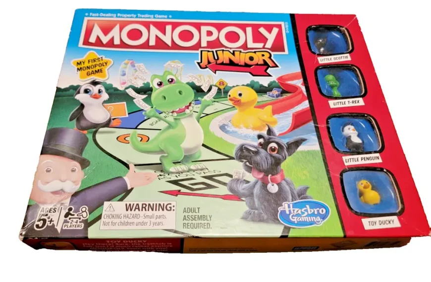 A box of "Monopoly Junior" board game by Hasbro Gaming, intended for ages 5 and up with a warning for a choking hazard. The cover features colorful cartoon illustrations of a dinosaur, a puffin, a dog, and other game pieces, alongside small windows displaying the actual game tokens: Little Scottie, Little T-Rex, Little Penguin, and Toy Ducky.