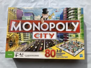 The image shows a "Monopoly City" board game box featuring the game's title in large letters, a 3D illustration with buildings, and the iconic Monopoly man with a golden key. The box indicates the game is for ages 8 and up, suitable for 2-6 players, requires 2 batteries (not included), and includes 80 3D buildings. The Hasbro logo is also visible.