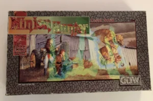 A board game box titled "Minion Hunter" with an image of two armed characters crouching near a tractor and facing a large, monstrous creature emerging from behind a building. The text indicates it is "A DARK CONSPIRACY Boardgame" by Lester Smith, suitable for ages 12 and up.