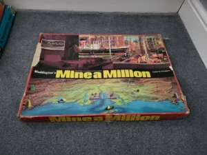 Vintage "Waddington's Mine a Million" board game box on a carpeted floor showing signs of wear with colorful graphics depicting mining and transportation scenes.