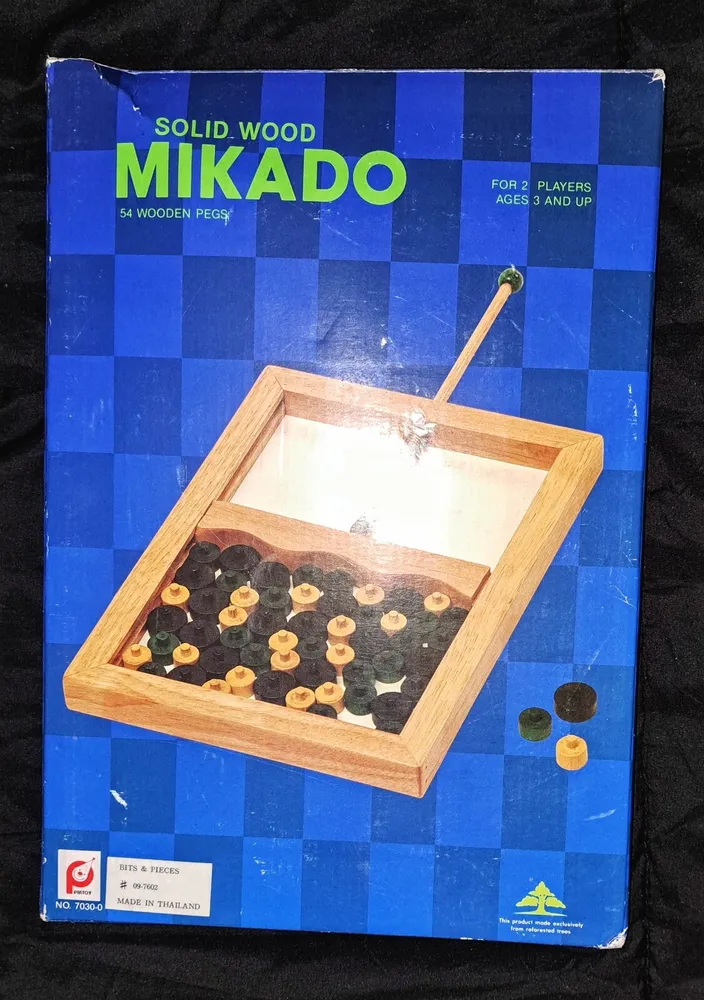 A worn-out board game box for "Solid Wood MIKADO," featuring 54 wooden pegs, intended for 2 players ages 3 and up. The box displays an image of the game contents, which include a wooden frame with pegs and checkered blue and white background. Visible logos indicate the product is from "Bits & Pieces" and "MADE IN THAILAND."