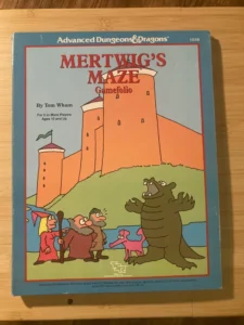 Alt text: The image displays the cover of a game called "Mertwig's Maze Gamefolio" for the Advanced Dungeons & Dragons series, by Tom Wham, intended for 2 or more players ages 10 and up. On the cover, colorful illustrations depict a whimsical cartoon scene with a surprised knight, a wizard, a pink creature, and a towering, friendly-looking dragon outside a large orange castle with towers. The TSR logo is visible, indicating the game's publisher. The game is placed on a wooden surface.