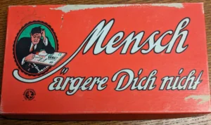 Vintage board game box with the title "Mensch ärgere Dich nicht!" in stylized white lettering on a red background; a graphic of a frustrated man holding a game board is featured in the upper left corner, with the logo of the manufacturer at the bottom left. The box shows signs of wear and paint chipping.